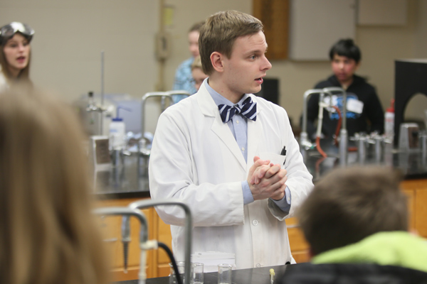 Student in lab coat and bowtie presenting to elementary and high school school students