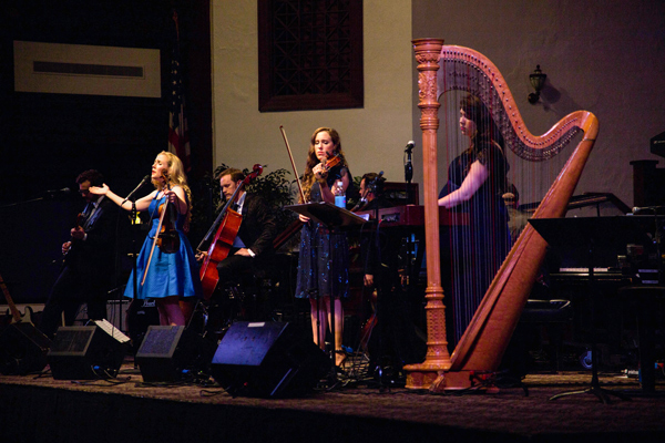 Musicians on stage singing, playing violin, harp, guitar, and cello