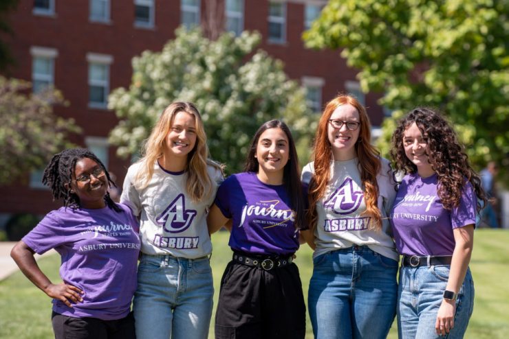 5 Asbury University students on campus wearing Asbury shirts and standing shoulder to shoulder posing for a photo while smiling with green grass and trees in the background