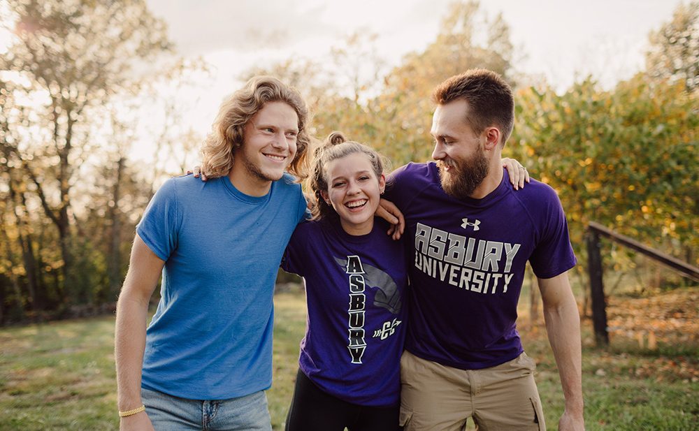 3 Asbury University students hug and smile with green grass and trees in the background while on campus