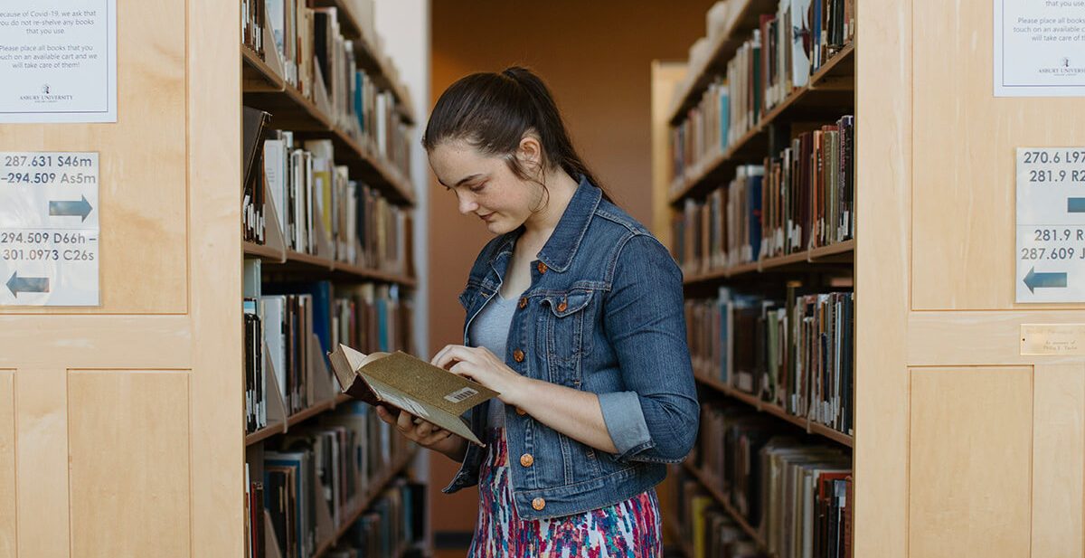 A female student reads a book between rows of books in the Asbury University library
