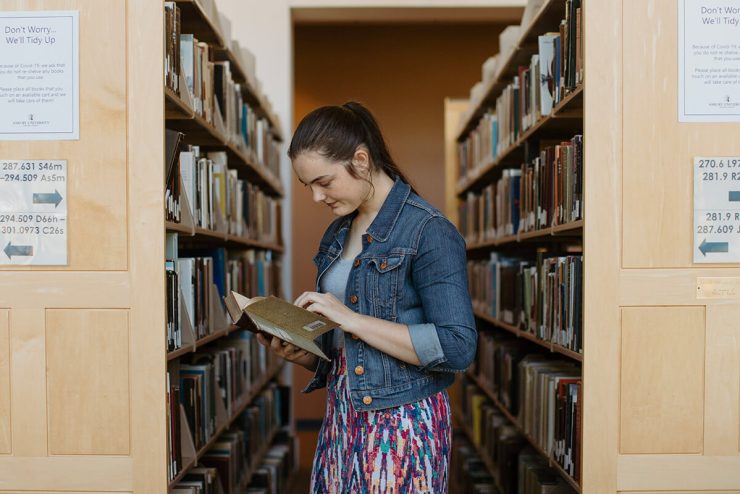 A female student reads a book between rows of books in the Asbury University library