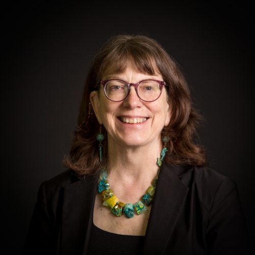 Dr. Slocum, a caucasian woman with dark auburn hair and purple glasses, wearing black with a turquoise necklace, against a black background.